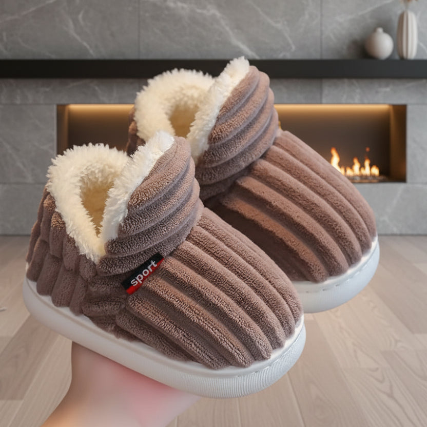 Pair of brown ribbed plush slippers with white fleece lining held up in a hand in front of a fireplace.