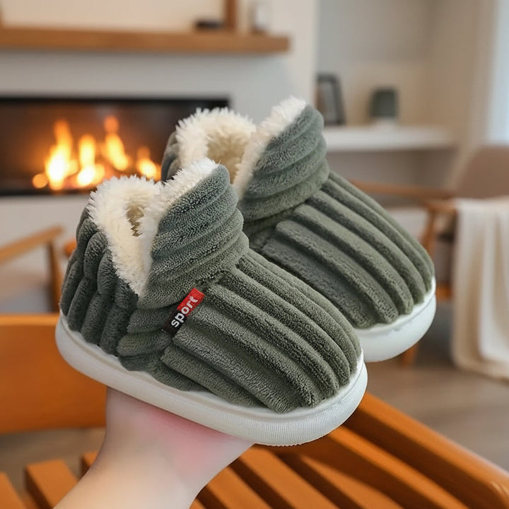 Pair of olive green ribbed slippers with white fleece collar held up in front of a lit fireplace.