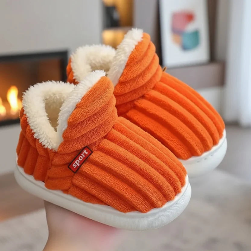 Pair of bright orange ribbed plush slippers with white fleece lining held in a hand beside a fireplace.