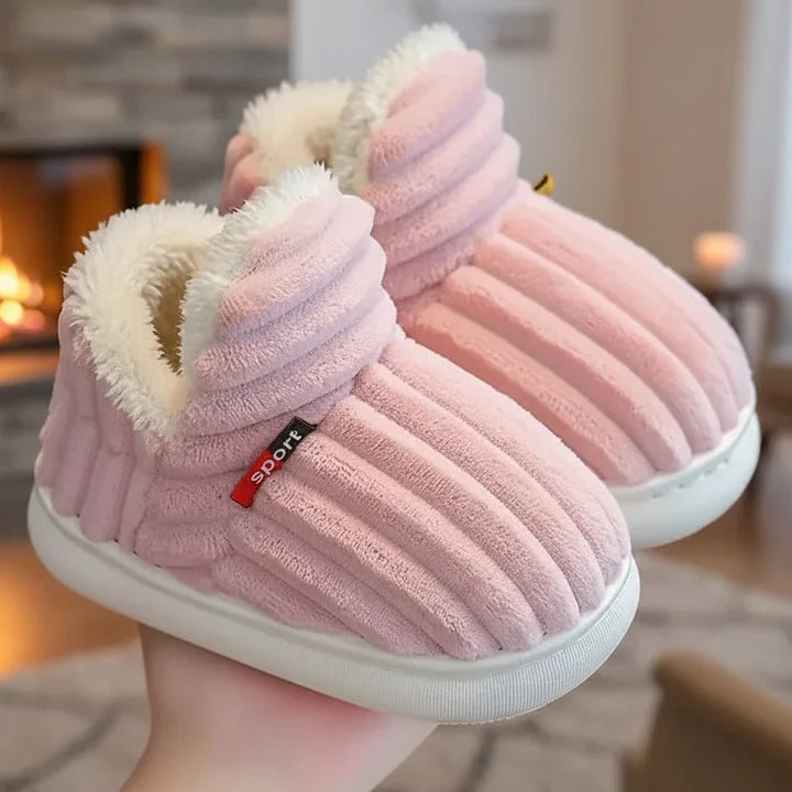 Pair of pink ribbed plush slippers with fleece lining held in a hand with a cozy fireplace in the background.