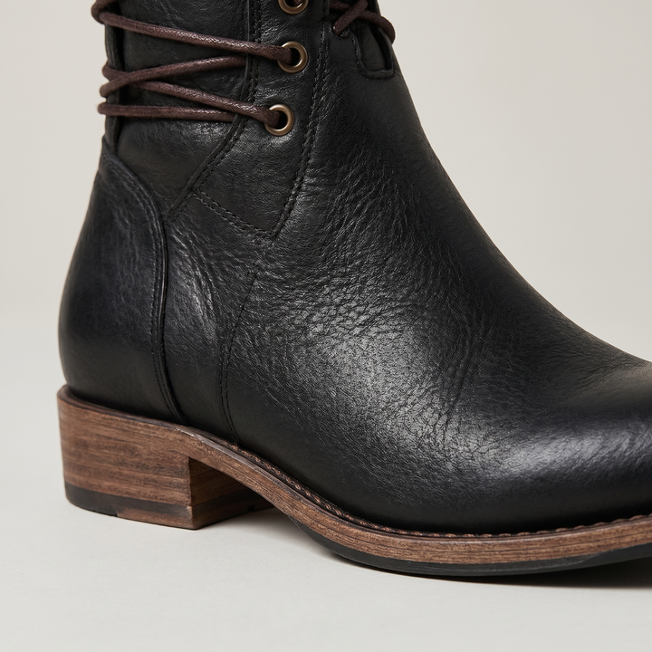 Close-up of a black boot's heel and side, showing grained leather, stitching, brass eyelets and the stacked heel.