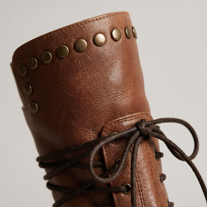Close-up of a tan boot's studded collar, grained leather texture and dark waxed laces tied in a knot.