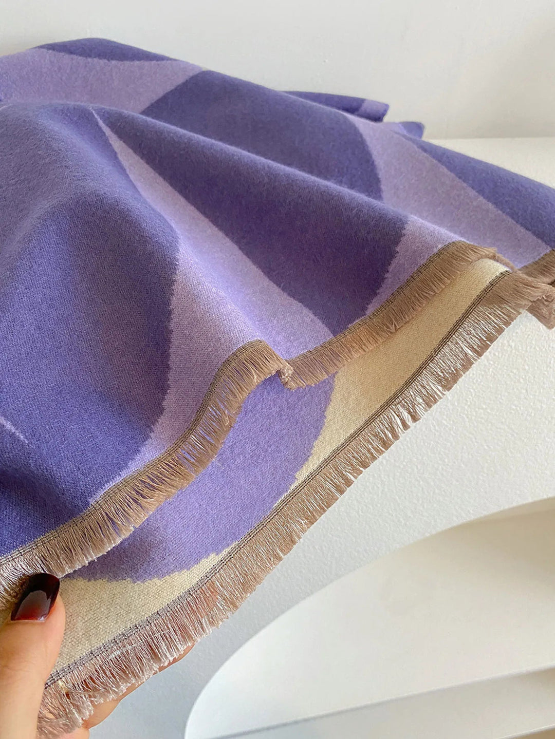 Close-up of the purple and cream scarf's fringed edge, held by a hand, showing the plush surface and tan fringe. 11