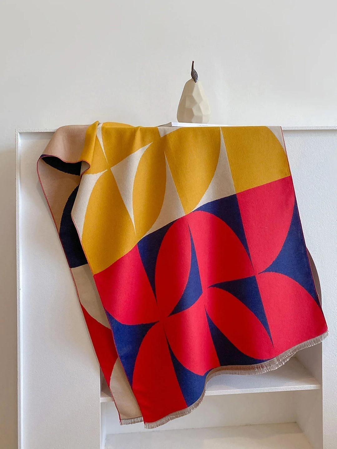 Red, mustard and navy scarf with a large geometric pattern hanging over a white shelf beside a pear-shaped ornament. 6