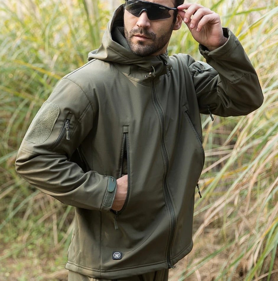Man in olive green jacket and sunglasses standing in tall grass, one hand in the pocket, the other adjusting his glasses. 11