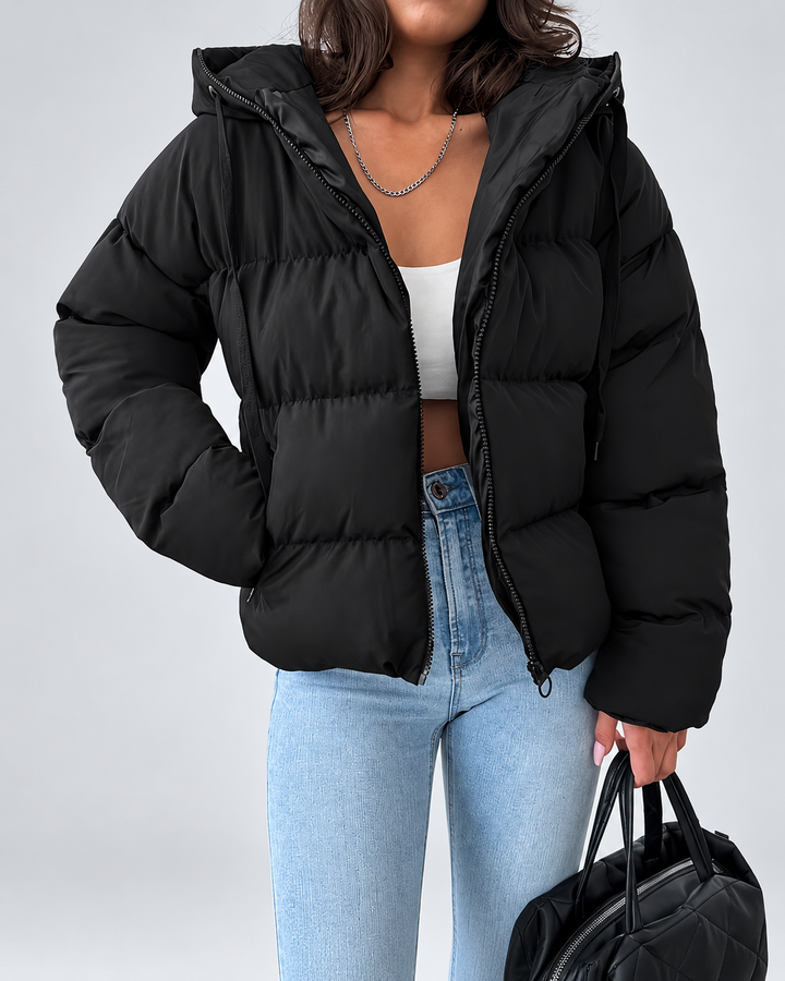 Model wearing the black puffer jacket unzipped over a white crop top and light blue jeans, holding a black quilted bag.